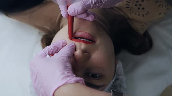 Closeup of a Permanent Makeup Master with a Red Pen Draws a Contour Sketch of Future Lips
