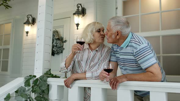 Senior Elderly Caucasian Couple Drinking Wine, Looking Ahead, Making a Kiss in Porch at Home alt