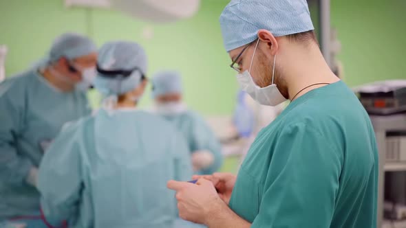 Assistant in Surgery Is Swiping Pages on Screen of Smartphone, Team of Surgeons Are Working alt