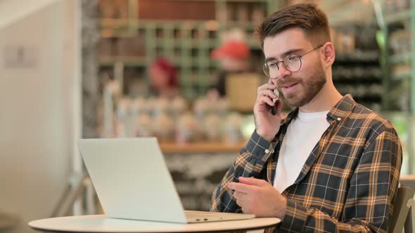 Young Man with Laptop Talking on Smartphone in Cafe  alt