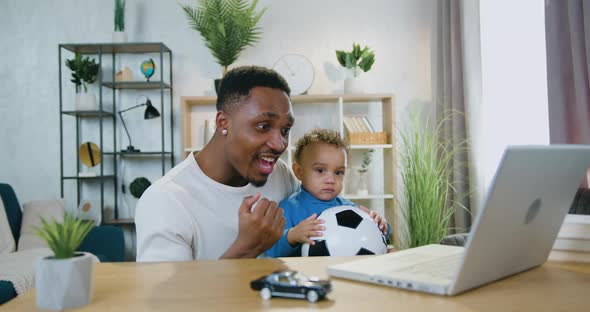 Dad Holding His Calm Baby Boy on Knees During watch Football Game on Computer alt