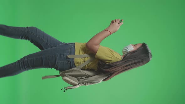 Girl Student Wearing A Mask And Playing Video Game On Mobile Phone While Walking On Green Screen alt
