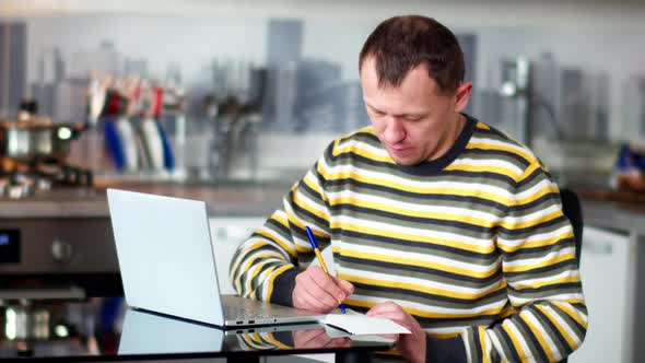 Young Man Working at Home Using Laptop, Making Notes in Notebook alt