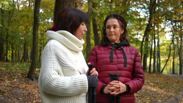 Thoughtful Focused Mature Woman Talking with Friend Standing in Autumn Park with Ski Poles alt