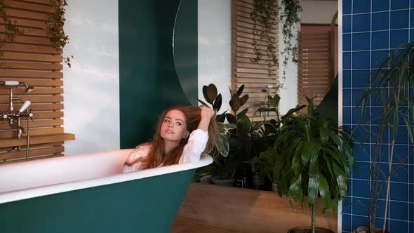 Pretty Girl With Long Hair In White Men's Shirt Sits In The Bathtub In The Bathroom alt