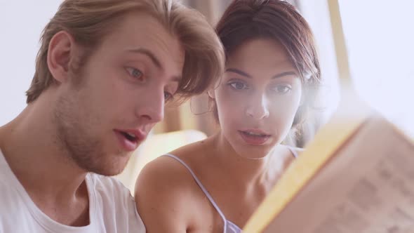 Young Beautiful Caucasian Pair Sitting in Bed and Reading Together Morning Magazine in Slowmotion alt