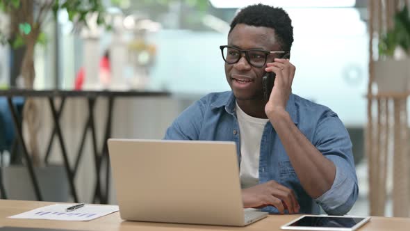 Young African Man Talking on Smartphone While Using Laptop alt