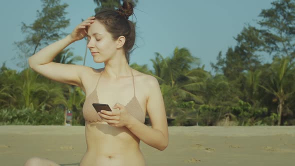 Sexy Girl in Bikini Holding Smartphone Reading Newsfeed on Tropical Beach Background. Young Woman
