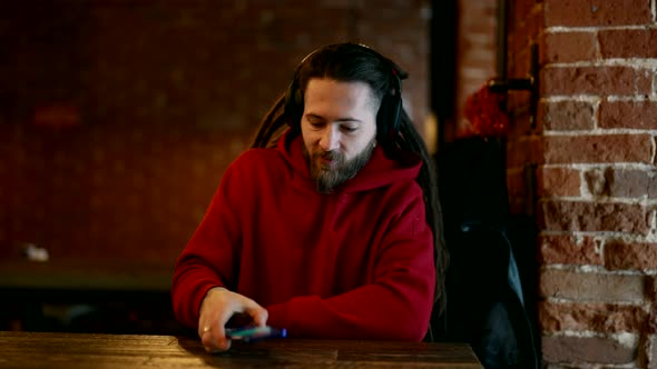 Hipster is Listening to Music By Headphones and Smartphone in Cafe Stylish Man with Dreadlocks alt