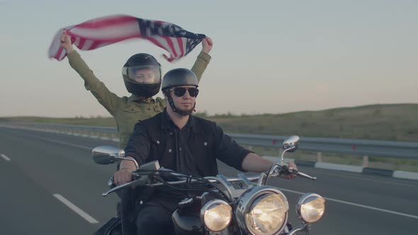 Man in Helmet and Black Glasses Rides with Woman Holding an American Flag Flying High in the Wind on alt