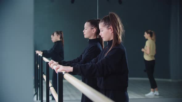 Little Girl Twins Synchronously Stretching By the Stand and Their Trainer Watching Them alt