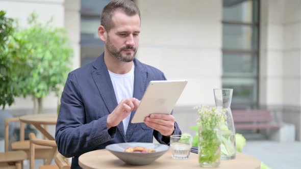 Man Browsing Internet on Tablet While Sitting in Outdoor Cafe alt