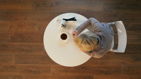 Young Attractive Business Woman Makes Notes in a Notebook Sitting in Cafe alt