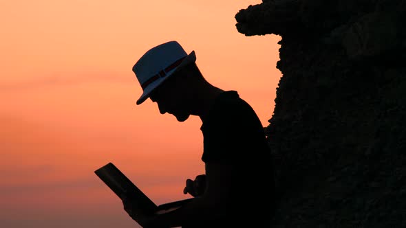 Attractive Young Man with Laptop Sitting on the Rock of Orange Sunset on Sea Shore. Happy Vacations