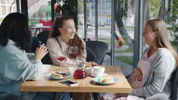 Cheerful Pregnant Woman Getting Gift Bag with Adorable Baby Shoes Talking to Friends in Cafe alt
