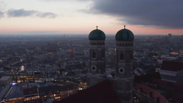 Flying Past Two Towers of Frauenkirche Cathedral in Munich, Germany at Dusk After Sunset with City alt