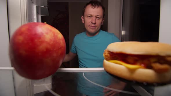 Man Opens the Refrigerator and Makes a Choice Between Healthy and Unhealthy Food, Apple or Burger