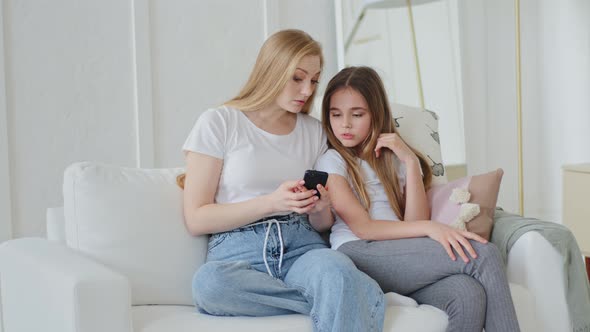 Concentrated Adult Mother and Teenage Daughter Child Girl Sitting at Home Couch Looking at Mobile alt