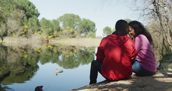 Smiling diverse couple embracing and sitting by lake in countryside alt