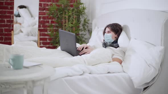 Wide Shot of Ill Tired Young Man in Covid Face Mask Lying in Bed Coughing and Using Video Chat on alt