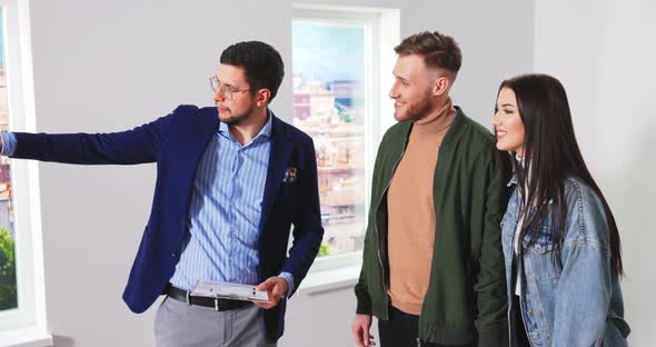 Cheerful Young Couple Standing in New Empty House Talking to Caucasian Male Real Estate Sales alt