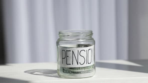 Male hand puts 100 dollars in a glass jar with the inscription pension. Concept of saving money.
