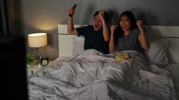 cheerful young couple watching sport TV and win a game with arm raised on a bed at night
