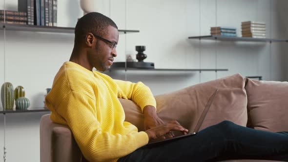 Young male in glasses typing on laptop keyboard sitting on sofa in home alt