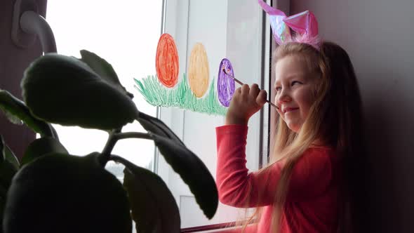Cute little girl in bunny ears painting Easter eggs with green spring grass on window at home alt