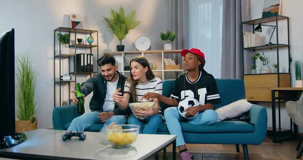 Mixed Race People Sitting in front of TV in Beautifully Decorated Room and Clinking Bottles alt