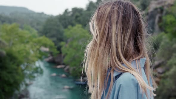 Side View of Woman Tourist Walks By the Road Bridge with View of Green Hills and River alt