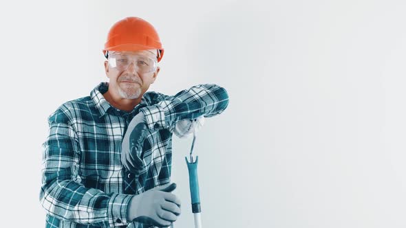 A Cheerful Grayhaired Man in a Hard Hat and Protective Glasses Holds a Roller with Paint in His Hand