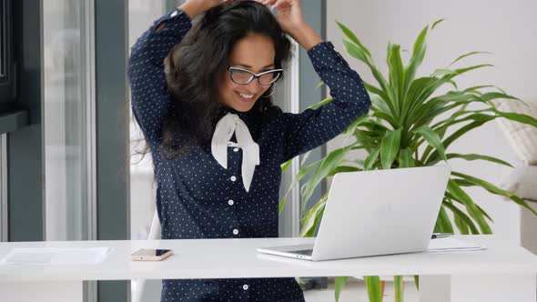 Active mixed race young woman freelancer student sits at desk in front of laptop, dances, moves wave