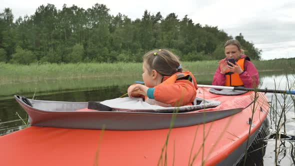 Little Girl Sits in Kayak with Mother Using Phone on Lake alt