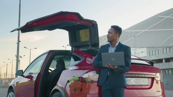 Close Up of African Man in Business Suit Standing Near Charging Electric Car and alt