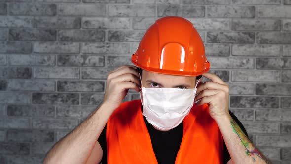 Male Construction Worker in Overalls Putting on Medical Mask on Face Background of Gray Brick Wall alt