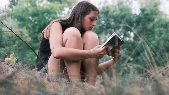 Young Woman Reads a Book Sitting on the Grass in Nature at Summer Day alt