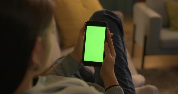 Young woman sitting on the couch at home and using her touch screen smartphone, green screen mock-up