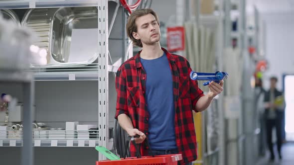 Concentrated Handsome Man Looking at Caulking Gun and Putting Tool in Shopping Basket alt