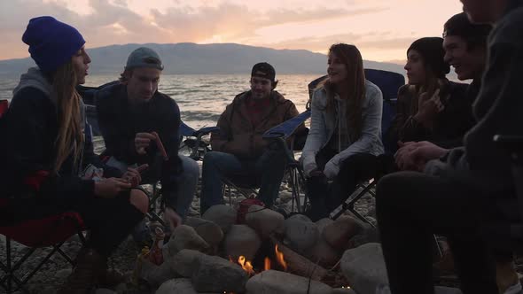 Group of millenials gathered around campfire at sunset alt