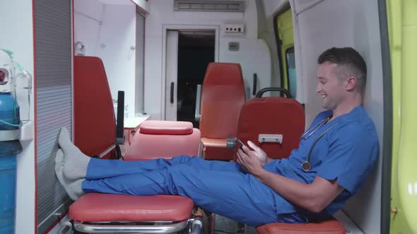 Man Sit with Phone in Ambulance Car, Watch Something and Smile. alt