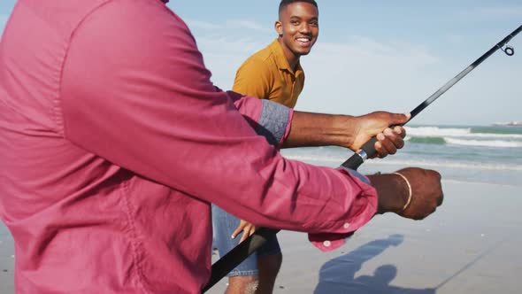 African american senior father and twin teenage sons standing on a beach fishing and talking alt