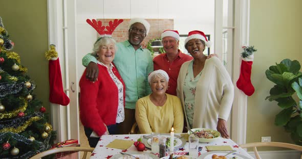 Portrait of diverse senior friends in christmas hats embracing and smiling at christmas dinner table alt